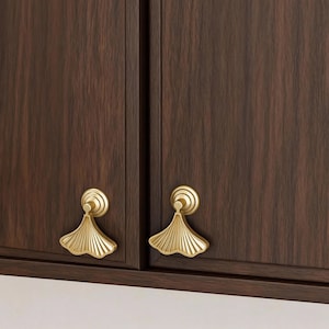 Könnte beinhalten: Nahaufnahme von zwei dunkelbraunen Schranktüren mit goldfarbenen, ginkgoblattförmigen Schrankgriffen. Die Griffe haben eine kreisförmige Basis und ein fächerförmiges Design, das den Schränken Eleganz verleiht.