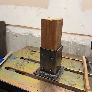 Könnte beinhalten: Ein Holzpfosten in einer geschweißten Stahlbasis. Der rechteckige, braune Pfosten passt in einen quadratischen Metallrahmen. Die Metallbasis hat eine quadratische Grundfläche mit vier Löchern. Das Bild wurde in einer Werkstatt aufgenommen.