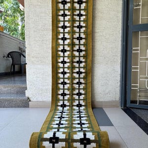 May include: A long, rectangular runner rug featuring a mustard yellow base with a repeating geometric pattern of white and black cross-shaped motifs. The design is framed by thin, dark green vertical lines running the length of the textile.