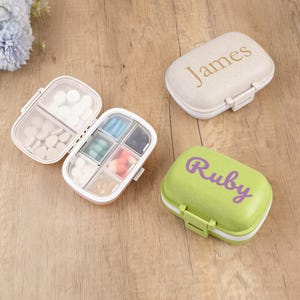 Könnte beinhalten: Drei personalisierte Pillenboxen. Eine ist geöffnet und zeigt Fächer mit Pillen. Die anderen beiden sind geschlossen, eine beige mit "James" und die andere grün mit "Ruby" in Schreibschrift. Auf einer Holzoberfläche.