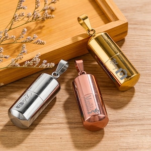 May include: Three metal pendant necklaces in gold, silver, and rose gold. Each cylindrical pendant has a screw-top lid and is engraved with a name and a small graphic. The pendants are displayed on a wooden tray.