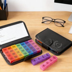 May include: A black leather pill organizer with a rainbow-colored pill case inside. Two additional pill cases are visible, one purple and one pink. A black leather journal with the name "Catherine" is also present, along with eyeglasses.