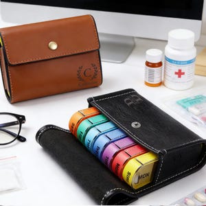 May include: A brown leather pill case with a gold button closure and a black leather pill organizer with a rainbow-colored weekly pill container. Also visible are prescription pill bottles and eyeglasses.