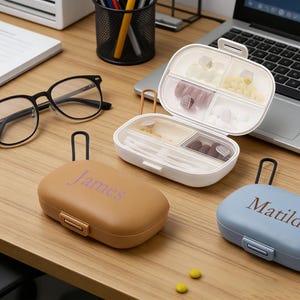 Könnte beinhalten: Drei Pillenboxen auf einem Holztisch. Eine ist geöffnet, gefüllt mit Pillen und Wattestäbchen. Die anderen beiden sind geschlossen, eine beige mit "James" und die andere blau mit "Matilda" bedruckt. Eine Brille mit schwarzem Rahmen und ein Laptop sind ebenfalls zu sehen.