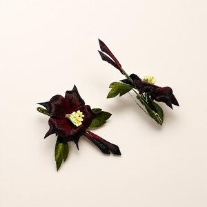 May include: Two floral hair accessories featuring deep burgundy, velvet-textured petals with pointed tips. Each piece includes a small cluster of pale yellow stamens at the center, green leaves, and a slender, dark stem-like extension attached to a clip.