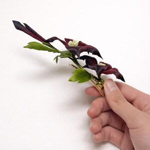 May include: A hair clip featuring two dark purple, petal-like flower shapes with light green centers, accented by several bright green leaves on a thin, gold-toned metal stem. The decorative floral piece is attached to a gold-colored metal alligator clip.