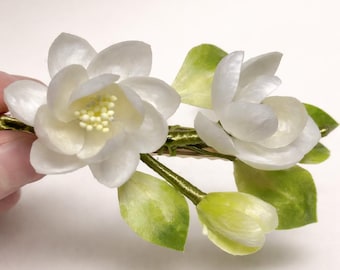 May include: A hair clip featuring two white flowers with yellow centers, accompanied by green leaves and a small bud. The floral elements are attached to a green stem-like base, creating a botanical design that mimics natural white blossoms.