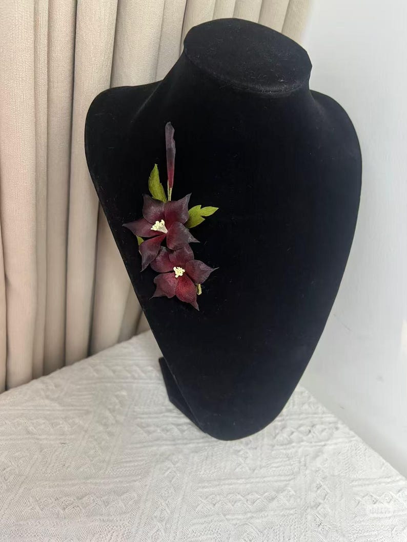 May include: A floral brooch featuring two dark burgundy, star-shaped flowers with white centers and a single slender, upright bud. The arrangement includes several small, textured green leaves attached to the stems, creating a botanical accessory.