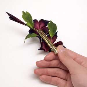 May include: A hair clip featuring a dark burgundy flower with deep purple undertones and several bright green leaves. The accessory is attached to a gold-toned metal alligator-style clip, which is visible on the back of the floral arrangement.