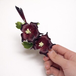 May include: A decorative floral accessory featuring two dark burgundy, star-shaped flowers with pointed petals and cream-colored centers. The piece includes small, vibrant green leaves and a slender, dark purple bud extending upwards from the base.