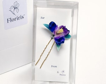 May include: A gold-toned metal hair pin featuring a decorative fabric flower with deep blue, purple, and light green petals and a yellow center. The pin is mounted on a white card labeled For and From, enclosed in a clear plastic display box.