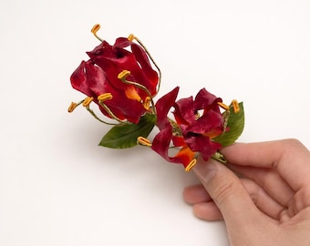 May include: A decorative floral accessory featuring two deep red, curled blossoms with vibrant orange centers and prominent yellow-tipped stamens. The flowers are accented by small, textured green leaves and thin, gold-toned wire stems.