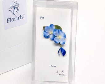 May include: A blue flower brooch with two main petals and a small bud, featuring white centers and green leaves, attached to a white card with the text For, From, and Floriris in a clear plastic display box with a white Floriris logo bag.