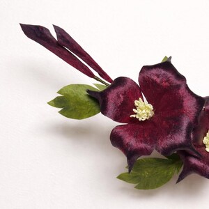 May include: A decorative floral arrangement featuring deep burgundy, velvet-textured petals with pointed tips. The flowers have prominent, pale yellow centers and are accented by small, serrated green leaves and slender, dark purple buds on stems.