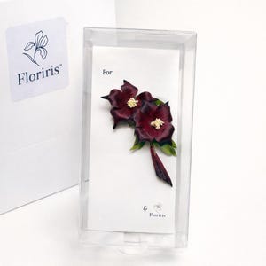 May include: A dark burgundy flower brooch with white centers and green leaves, presented on a white display card inside a clear plastic box. The card features the text "For" and the brand name "Floriris" with a small floral logo icon.