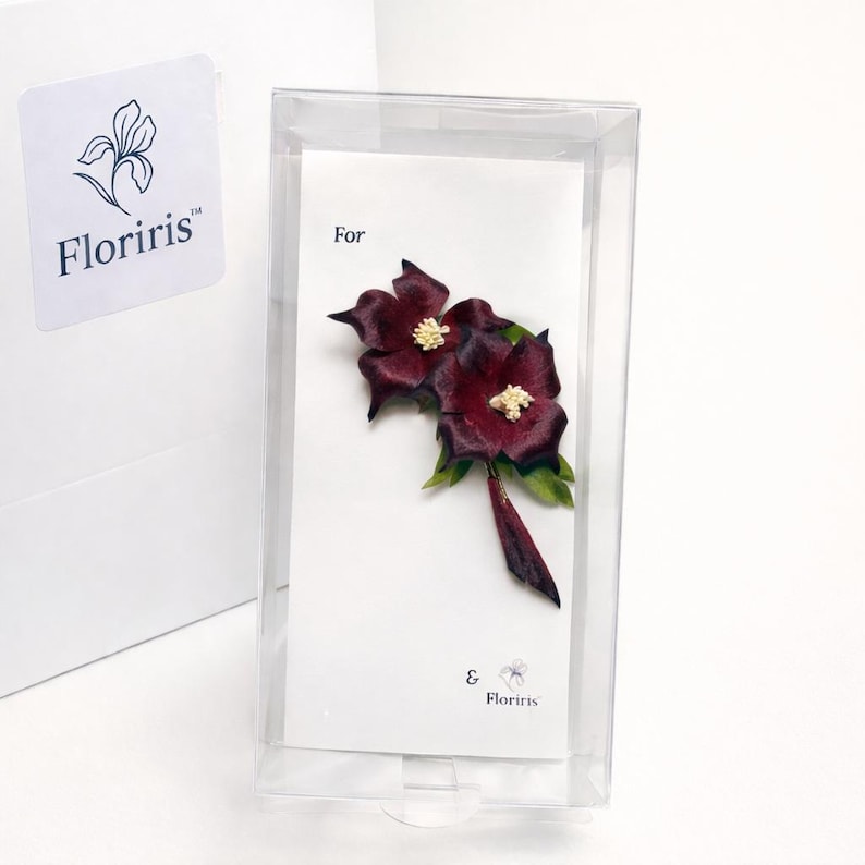 May include: A dark burgundy flower brooch with white centers and green leaves, presented on a white display card inside a clear plastic box. The card features the text "For" and the brand name "Floriris" with a small floral logo icon.