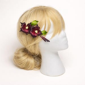 May include: A hair accessory featuring two deep burgundy, dark purple flowers with pointed petals and small yellow centers, accented by vibrant green leaves, attached to a stem-like base, displayed on a blonde mannequin head with a side bun.