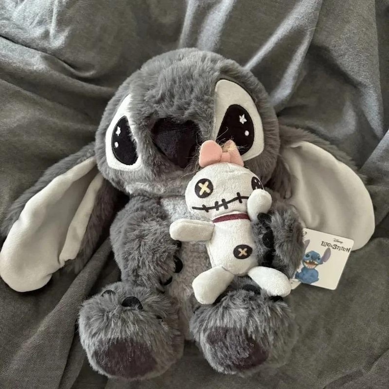 Puede incluir: Un peluche gris de Stitch con grandes ojos negros y una mu&ntilde;eca blanca m&aacute;s peque&ntilde;a con un lazo rosa. El mu&ntilde;eco de Stitch tiene orejas grandes y una textura esponjosa. Una etiqueta de Disney es visible.