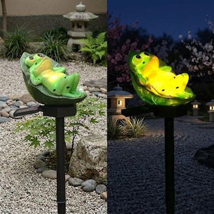 May include: A green and yellow frog statue reclines on a leaf, mounted on a black stake. The solar-powered garden ornament is set in a gravel bed with a Japanese-style stone lantern in the background. The frog glows at night.