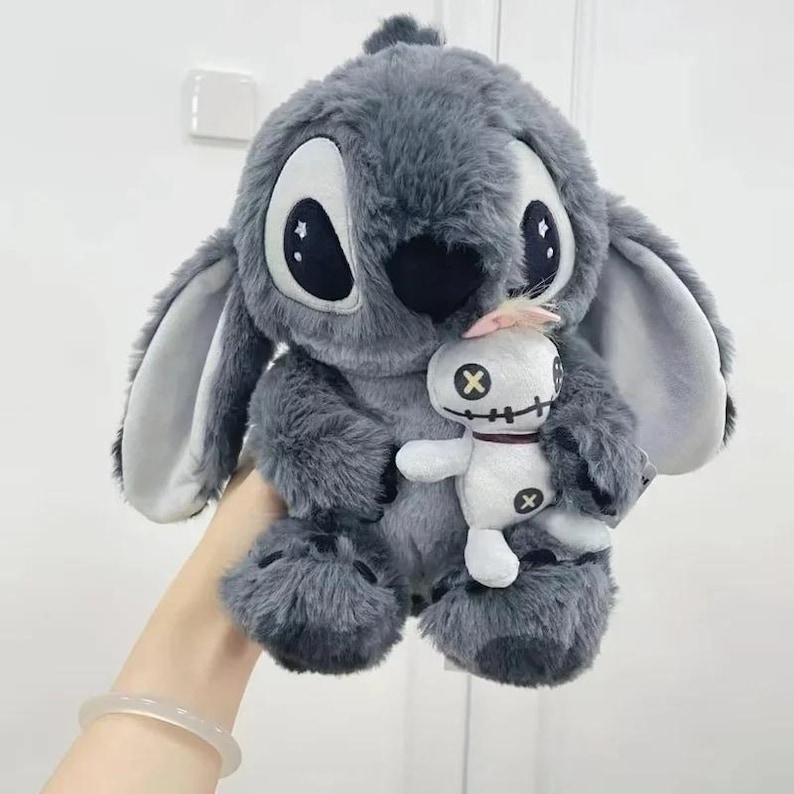Puede incluir: Un peluche gris con grandes ojos negros y orejas, sosteniendo una mu&ntilde;eca blanca m&aacute;s peque&ntilde;a. La mu&ntilde;eca tiene ojos negros en forma de cruz y un mech&oacute;n de pelo rosa. El peluche es suave y esponjoso.
