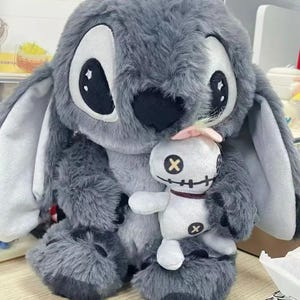 Puede incluir: Un peluche grande y gris de un personaje de dibujos animados con orejas grandes y ojos negros, sosteniendo una mu&ntilde;eca blanca m&aacute;s peque&ntilde;a con rasgos cosidos y un lazo rosa. El peluche tiene una textura esponjosa.