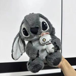 Puede incluir: Un peluche gris que se asemeja a un personaje de dibujos animados, con grandes ojos negros y orejas. Sostiene una mu&ntilde;eca blanca m&aacute;s peque&ntilde;a con una cara cosida y un lazo rosa. El peluche tiene una textura suave y peluda.