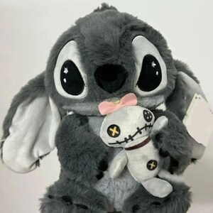 Puede incluir: Un peluche gris de Stitch sosteniendo una peque&ntilde;a mu&ntilde;eca vud&uacute; blanca con un lazo rosa. Stitch tiene ojos negros grandes, detalles blancos y orejas grandes. La mu&ntilde;eca vud&uacute; tiene detalles de punto de cruz negro y un collar marr&oacute;n. Una etiqueta en blanco est&aacute; adjunta.
