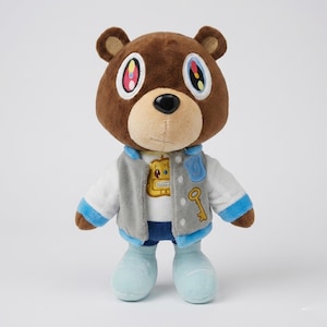 Puede incluir: Un oso de peluche marrón que lleva una chaqueta gris y blanca con detalles azules, una camisa blanca y zapatos azules. El oso tiene ojos coloridos y una nariz negra. Una llave dorada y un gráfico dorado están en la chaqueta.