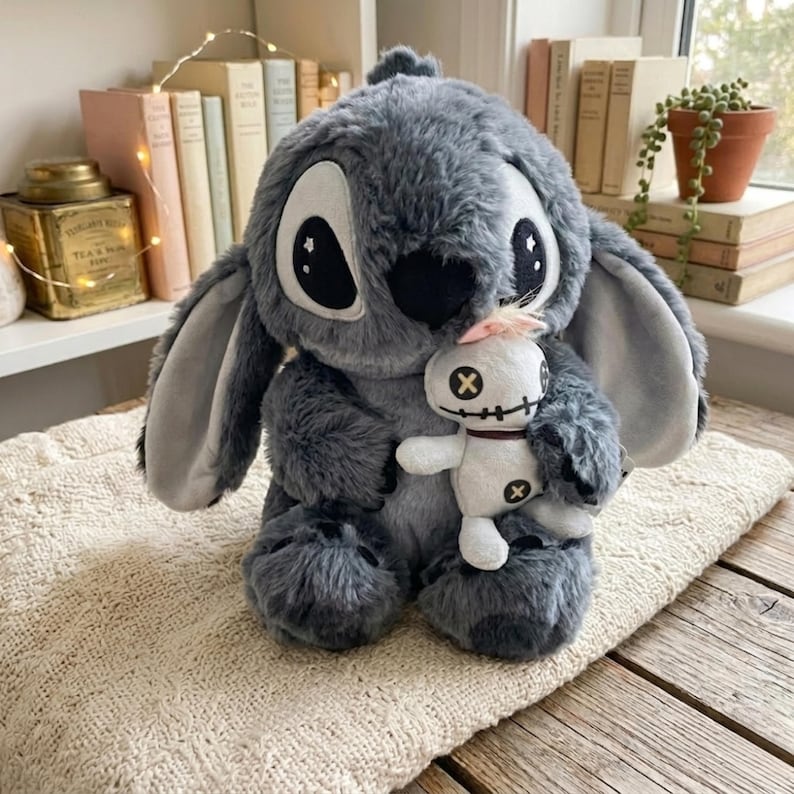 Puede incluir: Un peluche gris con orejas grandes y ojos negros, sosteniendo una mu&ntilde;eca blanca m&aacute;s peque&ntilde;a. El peluche tiene una apariencia cosida y est&aacute; sobre una superficie texturizada de color crema. La mu&ntilde;eca m&aacute;s peque&ntilde;a tiene una cara cosida y cruces.