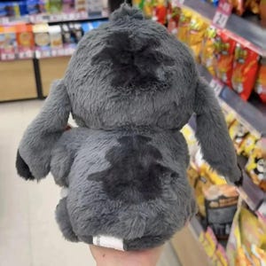 Puede incluir: Un peluche gris con forma de personaje de dibujos animados, visto desde atr&aacute;s. Tiene manchas grises m&aacute;s oscuras y detalles negros en las orejas y los pies. El juguete es suave y esponjoso, con una etiqueta visible en la parte inferior.