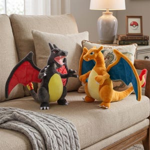 May include: Two plush Charizard figures, one black and one orange, are displayed on a beige sofa. The black Charizard has red wings and a yellow belly, while the orange one has blue wings. A gray cable-knit throw blanket is draped over the sofa.