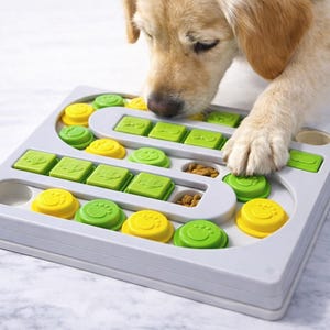 Pu&ograve; includere: Un giocattolo puzzle grigio per cani con pezzi scorrevoli gialli e verdi. Il giocattolo ha vari scomparti e coperchi scorrevoli. Un golden retriever interagisce con il giocattolo, usando la zampa e il naso per esplorare il puzzle.