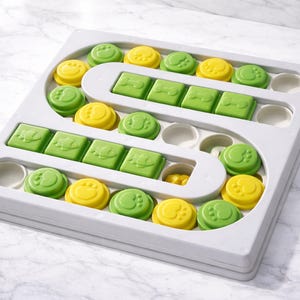 Pu&ograve; includere: Un puzzle per cani in plastica grigia con pezzi verdi e gialli. Il puzzle ha un design a labirinto con impronte di zampe e forme quadrate. I pezzi sono progettati per essere spostati per rivelare le prelibatezze.