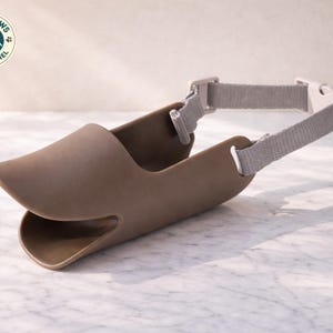 May include: A brown dog muzzle with a duckbill design, featuring a gray adjustable strap and buckle. The muzzle is made of a flexible material and is designed to fit comfortably. The Cozy Paws Pet Apparel logo is visible in the upper left corner.