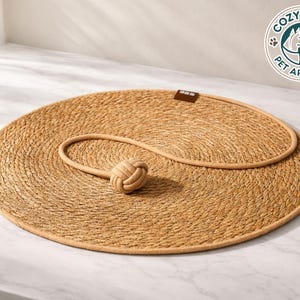 Könnte beinhalten: Ein runder, gewebter Haustier-Schlafplatz mit passendem Seilspielzeug. Das Bett hat eine hellbraune Farbe mit einem dunkleren Rand. Das Seilspielzeug hat einen geknoteten Ball an einem Ende. Das Logo von Cozy Paws Pet Apparel.