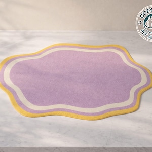 Könnte beinhalten: Ein lavendelfarbener Haustierteppich mit einer einzigartigen wolkenartigen Form. Der Teppich hat einen gelben Rand mit einem weißen Innenbesatz. Das Cozy Paws Pet Apparel-Logo ist sichtbar. Dieser Teppich ist für Haustiere konzipiert.