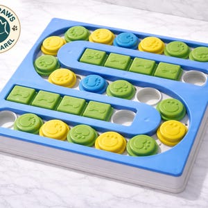 Pu&ograve; includere: Un giocattolo puzzle per cani blu e bianco con pezzi verdi, gialli e blu. Il giocattolo ha un design a labirinto con forme di ossa e impronte di zampe. Il logo Cozy Paws Pet Apparel si trova nell'angolo in alto a sinistra.