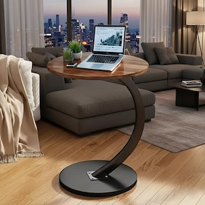 May include: A modern, round side table with a dark brown wooden top and a curved, black metal base. A laptop, small plant, and phone sit on the table. The table is in a living room with a city view.