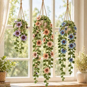 May include: Three hanging crochet planters with colorful flowers. Each planter is suspended by a natural rope. The flowers are in shades of purple, peach, and blue, with green foliage. The planters are set against a window.