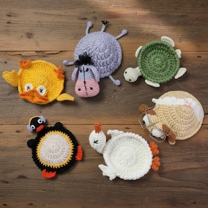 May include: A collection of handmade crochet coasters in various animal shapes, including a yellow duck, a purple hippo, a green turtle, a penguin, a sheep with glasses, and a white chicken. The coasters are arranged on a wooden surface.