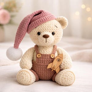May include: A handmade crochet teddy bear wearing a pink nightcap with a white pom-pom. The bear has a wooden rocket attached to its overalls. The bear is cream-colored with brown accents.