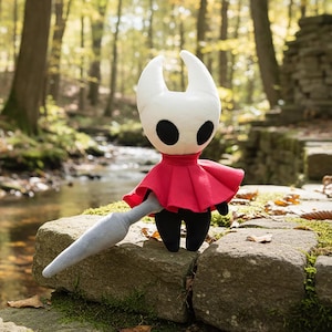 May include: A plush doll of a character with a white head, large black eyes, and a red cape. The doll has a gray, pointed weapon and black legs. It is posed on a stone surface with a forest background.