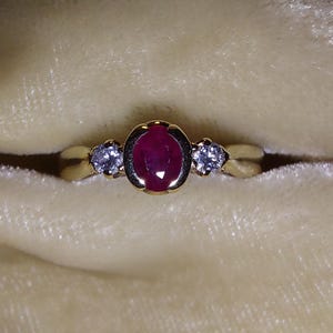 Peut inclure: Une bague en or ornée d'un grand rubis ovale au centre, flanqué de deux petites pierres précieuses claires et rondes. Le design de la bague est simple et élégant, mettant en valeur le rouge vif du rubis et l'éclat des pierres d'appoint.
