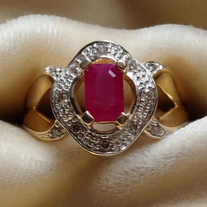 May include: A gold ring featuring a rectangular, deep red gemstone at its centre, encircled by a halo of small, sparkling diamonds. The ring has an ornate design with gold detailing.