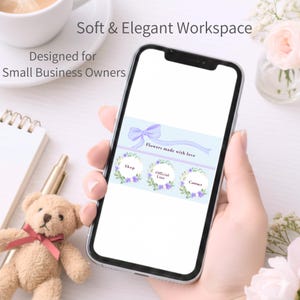 以下が含まれることがあります： スマートフォンには、「Flowers made with love」の文字と花の装飾が施された淡い青色のインターフェースが表示されています。画面には、「Shop」、「Official Line」、「Contact」とラベル付けされた3つの円形ボタンがあります。テディベアとコーヒーカップも写っています。