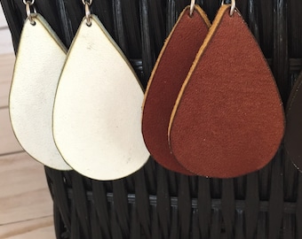 May include: Close-up of several pairs of teardrop-shaped earrings in various colours. The earrings are made of leather and have a smooth, textured surface. Colours include white, reddish-brown, and dark brown. The earrings are hanging from silver hooks.