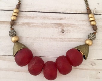 Puede incluir: Un collar con grandes cuentas rojas texturizadas, intercaladas con cuentas doradas y pequeños acentos brillantes. Tiene una cadena y detalles metálicos de color bronce, creando un estilo rústico y bohemio.