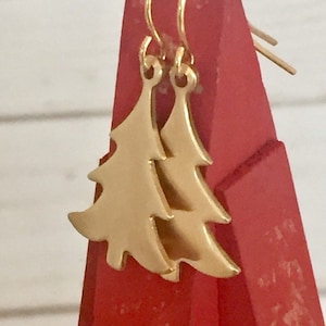 May include: A pair of gold-tone dangle earrings featuring a Christmas tree design. The earrings are on a red display stand.