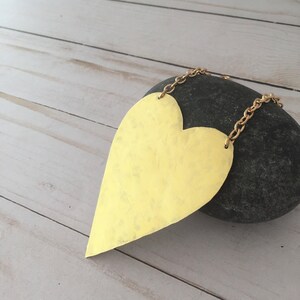 Op de afbeelding: Een grote, goudkleurige hartvormige ketting met een gehammerde textuur. De ketting hangt aan een gouden ketting en ligt op een grijze steen.