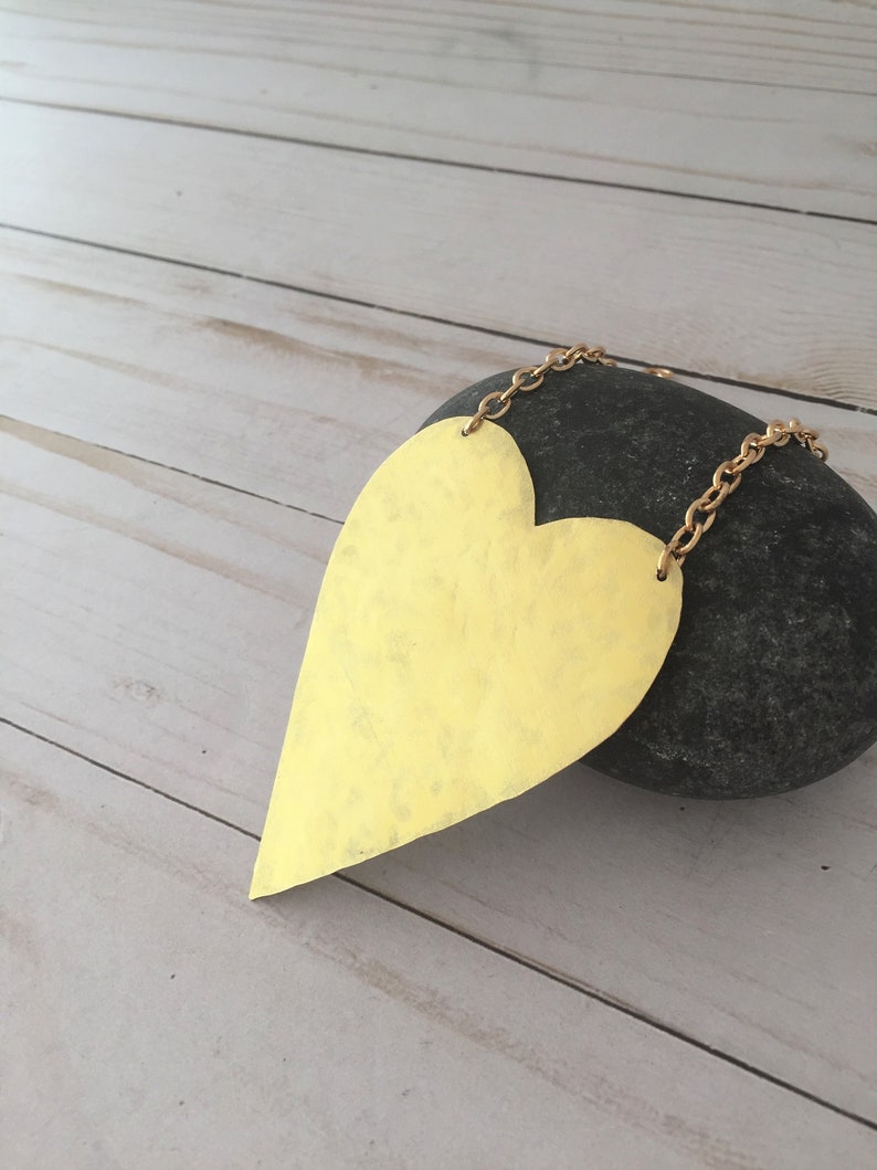 Op de afbeelding: Een grote, goudkleurige hartvormige ketting met een gehammerde textuur. De ketting hangt aan een gouden ketting en ligt op een grijze steen.